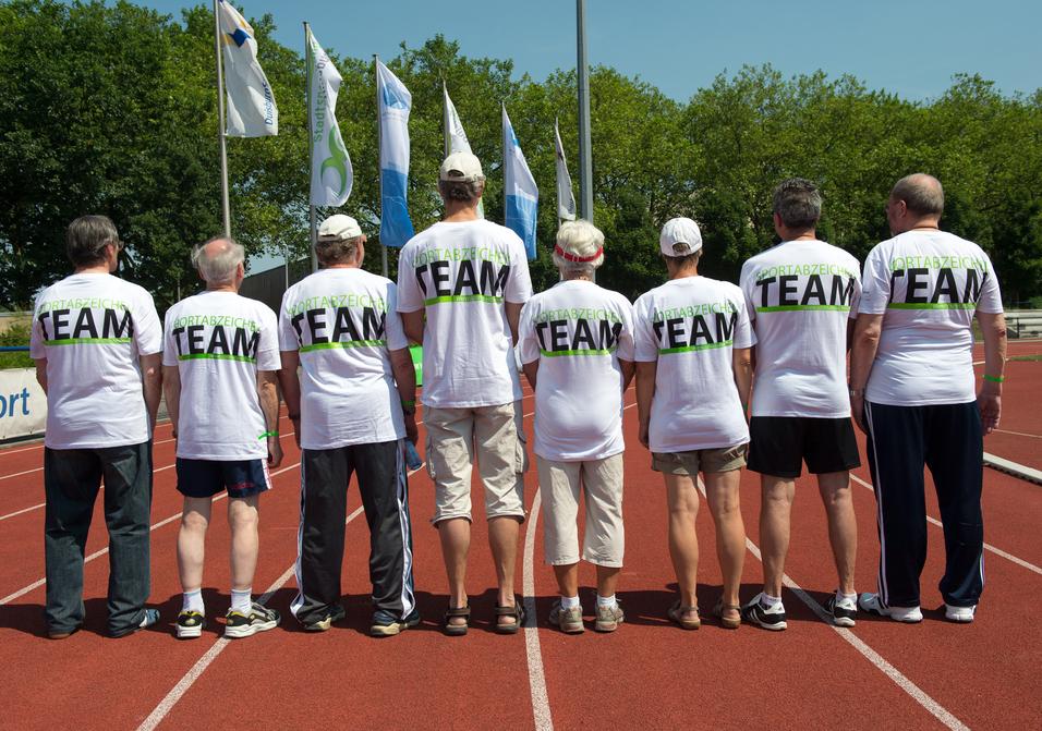 Gruppe von sieben Personen in weißen T-Shirts mit dem Aufdruck "TEAM" auf einer Laufbahn, Blick von hinten.